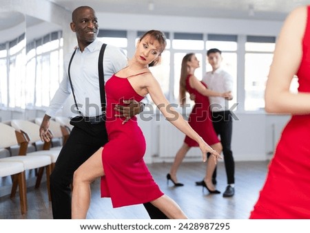 Elegant couple, attractive woman in red dress and stylish African American man, performing expressive tango in bright, modern dance studio during group class