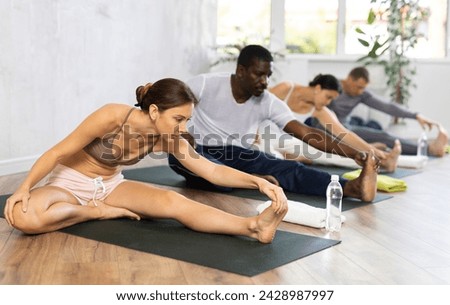 Portrait of young woman making stretching legs with multinational group sitting on sports mat and making yoga exercise at