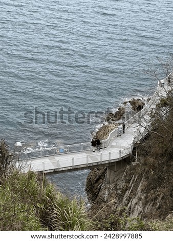 an elevated pedestrian walkway constructed along a rugged coastline. The walkway is supported by concrete beams, extending over the sea and hugging the natural contours of the cliffside Royalty-Free Stock Photo #2428997885