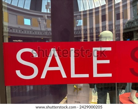 A vibrant red sale sign is prominently displayed in the window of a store. The sign features bold white lettering announcing a sale to passersby on the street outside. 