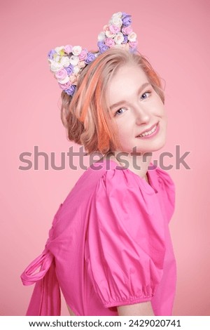 A cute blonde teenage girl with a short haircut poses in a pink dress and a lovely headband with floral kitty ears. Pink background. Kids and teenage fashion. Spring-summer look.