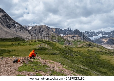 Photographer in vivid red wind jacket with hood and camera in hands in high mountains. Guy on stony hill takes pictures in scenic alpine valley with view to large glacier in sunlight under cloudy sky.