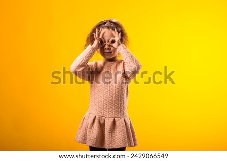 Portrait of smiling child girl looking through ok gesture over yellow background. Positive emotions concept