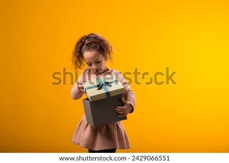 Portrait of smiling kid girl holding giftbox and looking inside the box over yellow background. Birthday and celebration concept