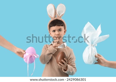 Thoughtful little boy wearing bunny ears with chocolate egg and female hands holding Easter gifts on blue background Royalty-Free Stock Photo #2429086301