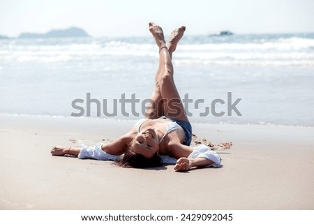 A beautiful girl with a wet hair and an Asian face type, a good slender figure posing on a sandy beach against the background of the ocean. Dressed jeans shorts and shirt