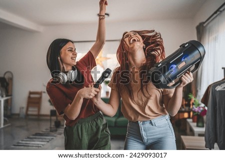 Two women young caucasian friends or sisters have fun at home females dance and sing karaoke hold microphone listen to the music happy smile joyful rhythm real people copy space