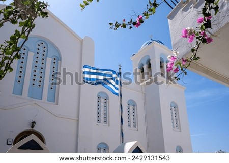 View of the greek flag waving in the air next to a bougainvillea and whitewashed church in the background in Ios Greece
