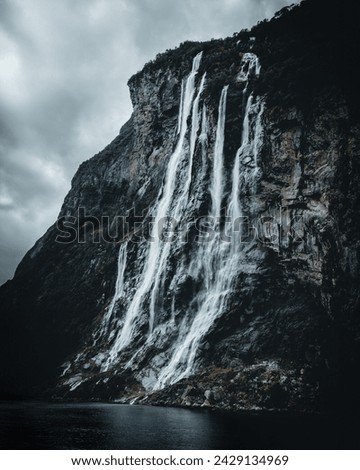 The Seven Sisters waterfall cascades with grace and power down the rugged cliff face, surrounded by the lush green embrace of the Norwegian fjords. Droplets catch the light, creating a misty veil that