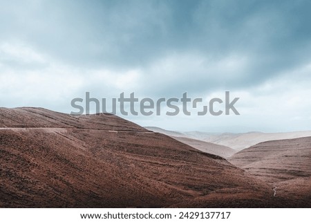 Layers of red and brown earth rise starkly against a dramatic sky, with a winding road cutting through the barren beauty of the tlas Mountains. The raw, rugged terrain speaks of the earth's deep histo Royalty-Free Stock Photo #2429137717