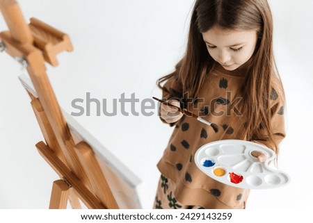 a 6-year-old girl with a brush in her hands in front of an easel with a canvas on an isolated white background, studio cyclorama