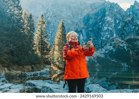 Rear shot of traveler 60s woman influencer in sportswear with backpack standing on the rocky shore and making selfie on smart phone. Taking photo of green hills and mountains on Morskie Oko lake