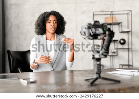 An African-American businesswoman engages confidently with a camera, gesturing as if explaining a complex concept during a video blog or webinar session in a modern office environment.