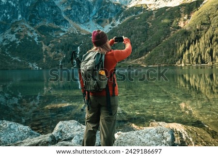 Rear shot of traveler woman in sportswear with backpack standing on the rocky shore and making selfie on smart phone. Taking photo of green hills and mountains on Morskie Oko lake. Hiking travel and