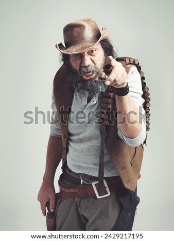 Senior cowboy, point and angry face at studio, costume and holster for western portrait looking scruffy. Old man, sheriff hat and dirty male for texas, grey background and annoyed gesture for outlaw Royalty-Free Stock Photo #2429217195
