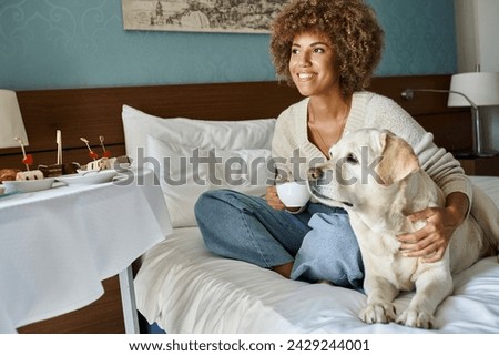 cheerful african american woman holding cup and cuddling labrador dog near room service breakfast Royalty-Free Stock Photo #2429244001