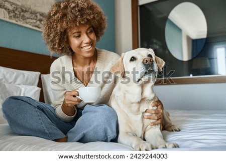happy african american woman holding cup of coffee and cuddling her labrador in pet friendly hotel Royalty-Free Stock Photo #2429244003