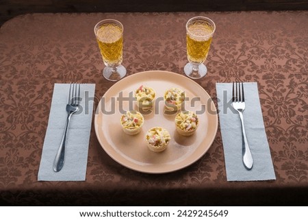 Sparkling champagne in two glasses next to a plate with salad in tartlets on a brown tablecloth and forks. Horizontal photo.