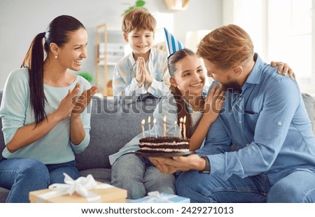 Happy girl celebrating birthday with family sitting on sofa in the living room at home. Smiling father congratulating daughter giving her cake at party. Family holiday celebration concept.