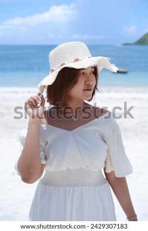 Portrait of young smiling Asian girl with white sand beach and clear blue sea background. Teenage girl wearing sunglasses, straw sun hat, white dress enjoying holidays. Outdoor summer travel concept, 