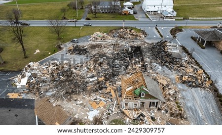 An Aerial View Of A Devastated Building Amidst Intact Structures With Cars On The Road And Green Fields In The Background Under A Cloudy Sky. Royalty-Free Stock Photo #2429309757