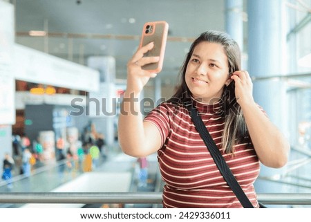 Selfie, young woman in the airport hallway next to the window ready to travel, with a big smile. Smartphone for taking a profile picture on social media app before boarding the airplane, while fixing 