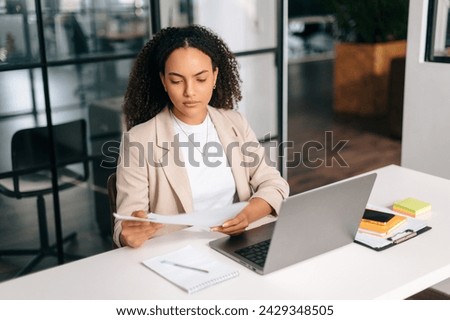 Serious confident brazilian or hispanic curly haired business woman, company ceo, financial manager, sitting at a work desk in modern office, working in a laptop, focused studying financial documents Royalty-Free Stock Photo #2429348505