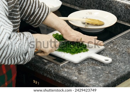 Close-up of an elderly woman's hands as she finely chops fresh green parsley on a white cutting board in a home kitchen