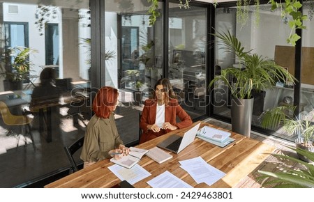 Two busy happy business women of young and middle age talking in green office sitting at desk. Professional ladies employee and manager having conversation using laptop at work. Candid top view. Royalty-Free Stock Photo #2429463801
