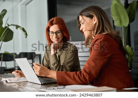 Two happy busy female employees working together using computer planning project. Middle aged professional business woman consulting teaching young employee looking at laptop sitting at desk in office Royalty-Free Stock Photo #2429463807