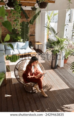 Busy elegant mature middle aged professional business woman wearing suit holding digital tablet using tab working sitting in comfortable chair in sunny office with green plants. Vertical top view. Royalty-Free Stock Photo #2429463835