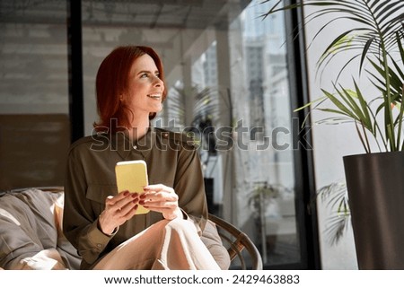 Happy smiling young professional business woman with red hair holding mobile cell phone in hands at work sitting in chair in sunny office with cellphone looking away at copy space using smartphone.
