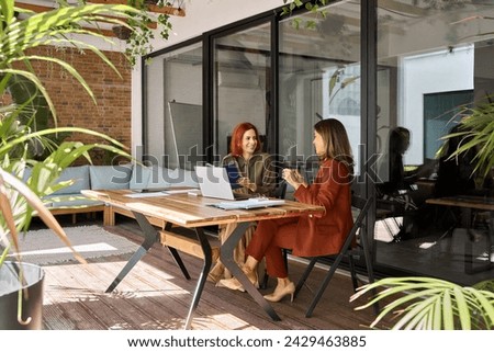 Two happy busy business women of young and middle age talking in green cozy office sitting at desk. Professional ladies executives having conversation sharing ideas using laptop at work. Candid shot. Royalty-Free Stock Photo #2429463885