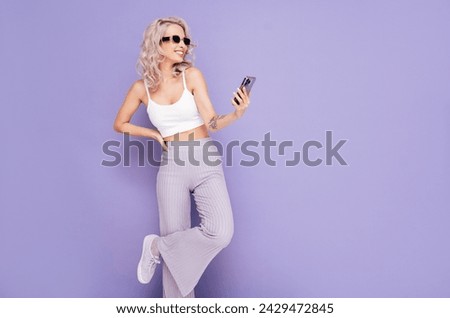 Young beautiful smiling female in trendy summer white top and pants clothes. Carefree blond woman posing near violet wall in studio. Positive model holds smartphone, uses phone apps, looks at screen 