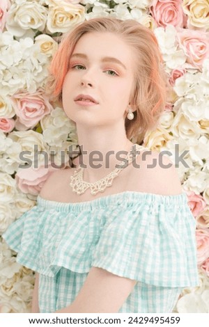 Beauty, cosmetics. A cute blonde teenage girl with a short haircut in a summer sundress and a pearl necklace poses against a wall of white and pink flowers. Delicate spring-summer look.