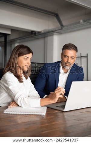 Team of diverse partners sitting at table mature Latin business man and European business woman discussing project on laptop in office. Two colleagues of professional business people working, vertical Royalty-Free Stock Photo #2429541937