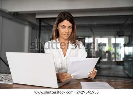 Focused European Latin business woman accountant holding documents, work at laptop pc doing online data base trade market analysis. Portrait 40s mature hispanic businesswoman with paperwork in office Royalty-Free Stock Photo #2429541955