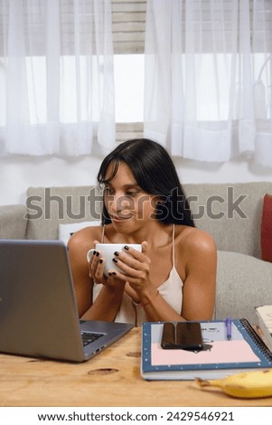 Vertical image of young happy woman at home sitting using laptop and drinking coffee, looking at screen happy working on her digital business.
