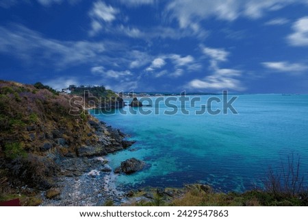 A rocky shore with a body of water and a blue sky with clouds, a body of water surrounded by a lush green hillside  Royalty-Free Stock Photo #2429547863