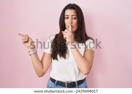 Young brunette woman standing over pink background asking to be quiet with finger on lips pointing with hand to the side. silence and secret concept. 