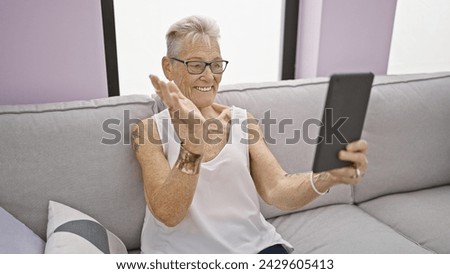Joyful, grey-haired senior woman having a fun video call sitting and relaxing on the sofa at home, expressing happiness with a warm smile