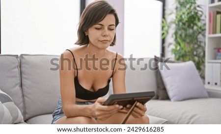 Emotive portrait, young beautiful hispanic woman, with short hairstyle, engrossed in a cherished framed photo, sitting on the sofa at home, expressing serious yet relaxed concentration.