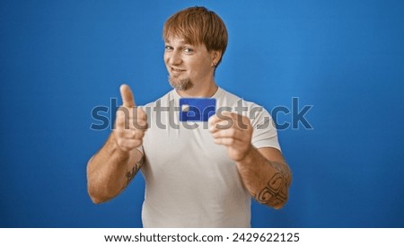 A tattooed young man with a beard poses in front of a blue wall, giving a thumbs up while holding a credit card.