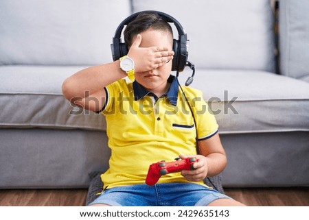 Young hispanic kid playing video game holding controller wearing headphones covering eyes with hand, looking serious and sad. sightless, hiding and rejection concept  Royalty-Free Stock Photo #2429635143