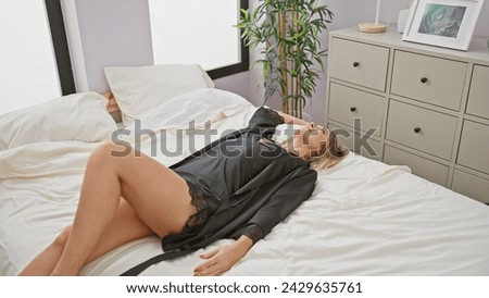 Portrait of a relaxed blonde woman in a black robe lying on a white bed in a modern bedroom