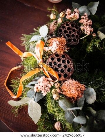 An overhead view of an advent wreath showcasing an array of textures with dried lotus pods, delicate pink blooms, eucalyptus leaves, and a satin orange ribbon.