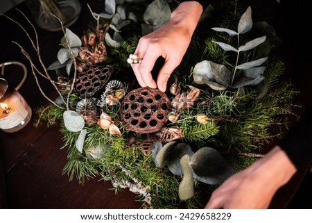 A person's hand delicately arranges a lotus pod within a lush advent wreath, surrounded by eucalyptus, pine, and warm candlelight Royalty-Free Stock Photo #2429658229