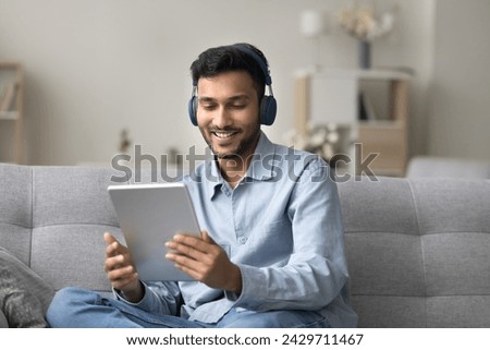 Young Indian man in modern wireless headphones sit on sofa watch new vlog or music videos on digital tablet, enjoy videocall talk with family, choose audio book spend pleasant time on internet at home Royalty-Free Stock Photo #2429711467