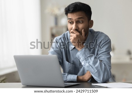 Bewildered confused or surprised Indian man sit at desk looking at laptop screen read news, experiences lack of understanding, feels confused, has problems with new apps or completing creative task Royalty-Free Stock Photo #2429711509