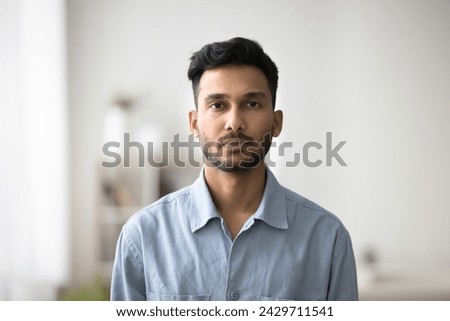 Serious Indian guy in casual blue shirt posing for camera standing alone in modern living room. Portrait of unsmiling millennial generation man, single homeowner or tenant head shot, profile picture Royalty-Free Stock Photo #2429711541